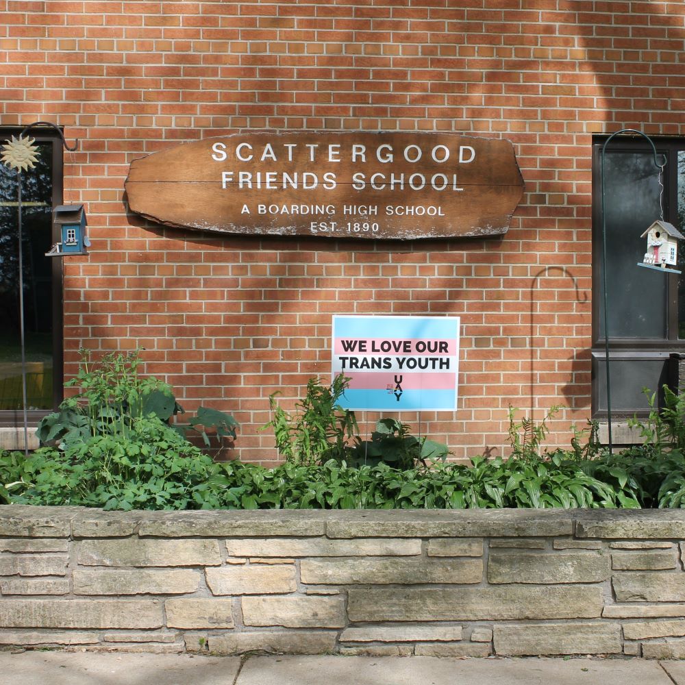 Photo of the main building and the Scattergood Friends School sign. In the garden is a yard sign reading "We love our trans youth"